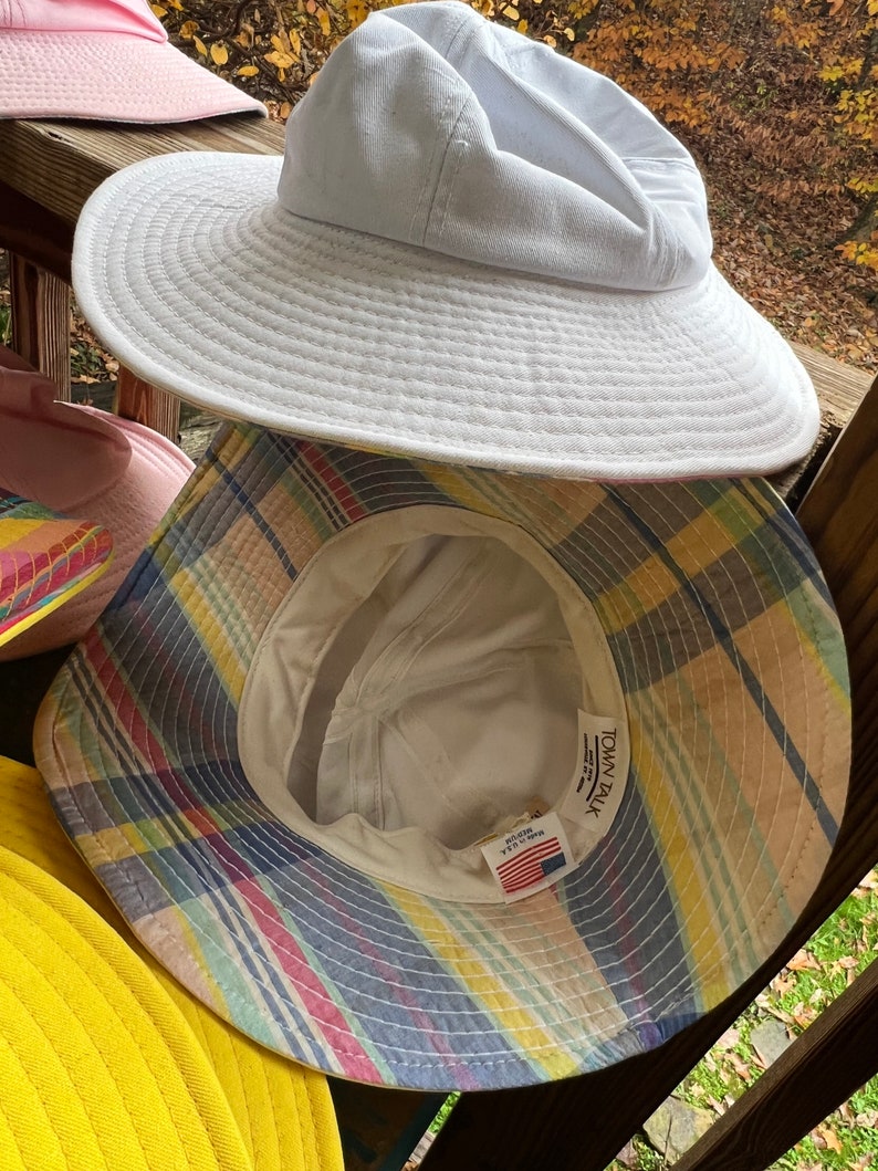 May include: A selection of sun hats in different colours. The top hat is white with a wide brim. Beneath it is a plaid bucket hat with yellow, blue, and pink stripes. Other hats in pink and yellow are also visible.