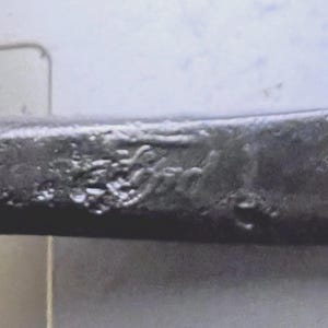 Puede incluir: Primer plano de un objeto metálico oscuro y texturizado con la palabra "Ford" grabada en su superficie. El objeto parece ser parte de una estructura más grande, posiblemente una herramienta o un componente de maquinaria.