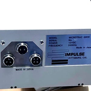 May include: A white electronic device with three circular ports on the left and a label on the right. The label reads: MODEL MICROTRAC 6800, SERIAL No., POWER 12VDC, FREQUENCY 200KHz, Made in Japan, IMPULSE PITTSBURG, CAL. The text MADE IN JAPAN is also printed below the ports.