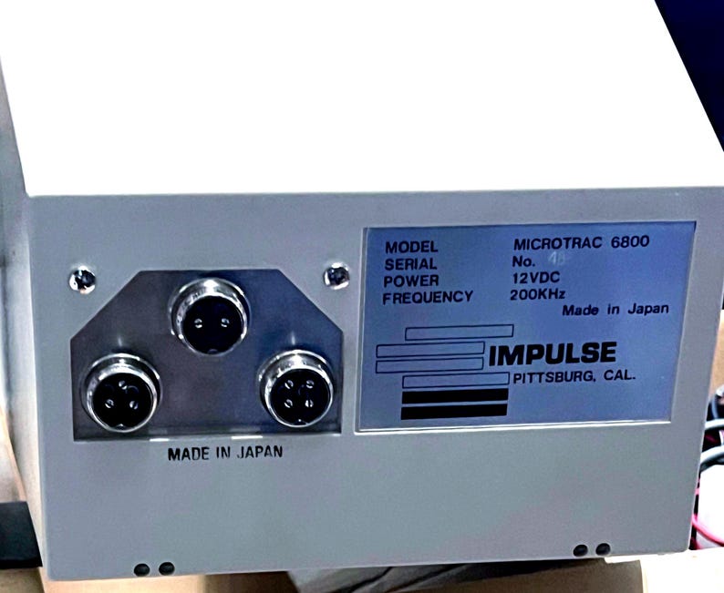 May include: A white electronic device with three circular ports on the left and a label on the right. The label reads: MODEL MICROTRAC 6800, SERIAL No., POWER 12VDC, FREQUENCY 200KHz, Made in Japan, IMPULSE PITTSBURG, CAL. The text MADE IN JAPAN is also printed below the ports.