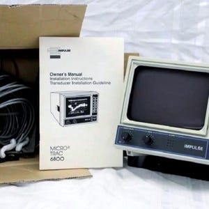 May include: An Impulse Micro-Trac 6800 marine fish finder unit with a gray casing, a black screen, and a control panel with buttons and knobs. Included are a coiled gray transducer cable, an owner's manual, and a cardboard shipping box.