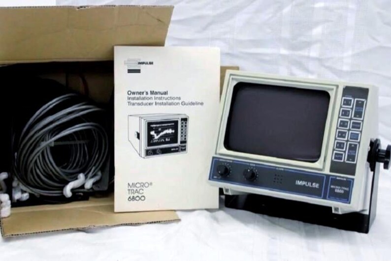 May include: An Impulse Micro-Trac 6800 marine fish finder unit with a gray casing, a black screen, and a control panel with buttons and knobs. Included are a coiled gray transducer cable, an owner's manual, and a cardboard shipping box.