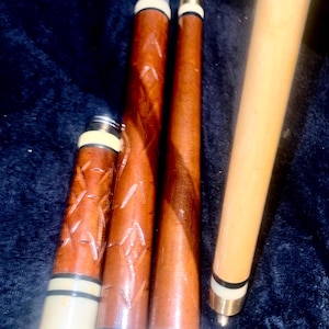 May include: A collection of pool cues and cue parts. The cues are made of wood, with some featuring decorative carvings. The tips are white and black. The cues are in various stages of assembly, with some pieces separate.