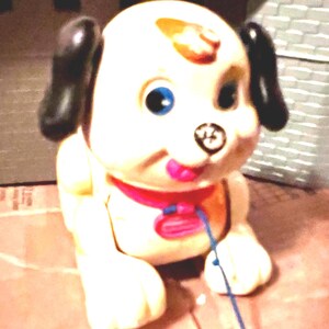 May include: A plastic toy dog with a cream-colored body, black ears, and blue eyes. The dog features a brown spot on its forehead, a black nose, and a pink tongue. It wears a red collar with a pink tag and has a thin blue string attached to the front.