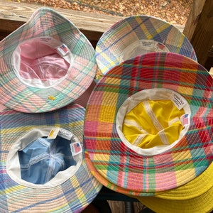 May include: Several plaid bucket hats in shades of pink, blue, yellow, and red. Each hat has a white interior lining and a "TOWN TALK" tag. The hats are displayed on a wooden surface, showcasing their colourful patterns.