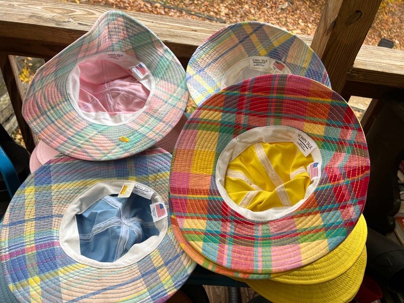 May include: Several plaid bucket hats in shades of pink, blue, yellow, and red. Each hat has a white interior lining and a "TOWN TALK" tag. The hats are displayed on a wooden surface, showcasing their colourful patterns.