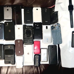 May include: A collection of various smartphones and a smartwatch, in different colors and designs. Some phones have cracked screens or missing parts. The devices are arranged on a dark surface, showcasing a variety of models.