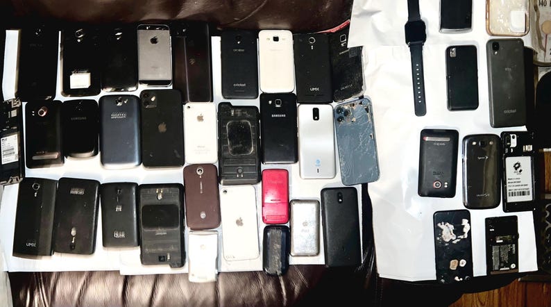 May include: A collection of various smartphones and a smartwatch, in different colors and designs. Some phones have cracked screens or missing parts. The devices are arranged on a dark surface, showcasing a variety of models.