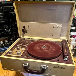 May include: A vintage Silvertone portable record player in a cream-coloured suitcase-style case. The device features a deep maroon turntable platter, a matching maroon tonearm, and a control panel with three knobs and a radio frequency display.