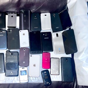 May include: A collection of various smartphones and a smartwatch, in different colors and conditions. Some phones have cracked screens. The devices are arranged on a dark surface, showcasing a variety of models and designs.