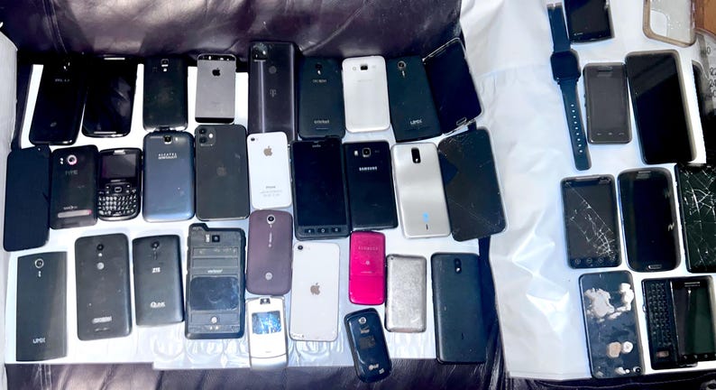 May include: A collection of various smartphones and a smartwatch, in different colors and conditions. Some phones have cracked screens. The devices are arranged on a dark surface, showcasing a variety of models and designs.