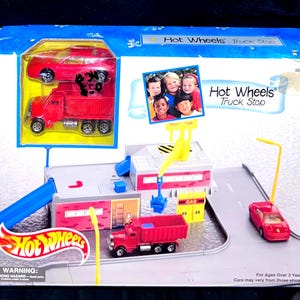 Może przedstawiać: Zestaw do zabawy Hot Wheels Truck Stop w oryginalnym pudełku. Zestaw zawiera czerwoną ciężarówkę i samochód, stację benzynową i niebieską rampę. Pudełko zawiera logo Hot Wheels i napis "Truck Stop".