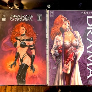 May include: Two comic book covers featuring female characters. The cover on the left is for "CYNDER #1" and features a woman with red hair wearing a black leather outfit. The cover on the right is for "SIRIUS DRAMA" and features a woman with red hair wearing a white dress and a red corset.