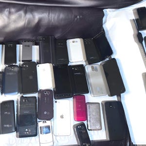 May include: A collection of various smartphones and a smartwatch, in different colors and designs. The phones are mostly black, with some white and other colored devices. Some phones have cracked screens.