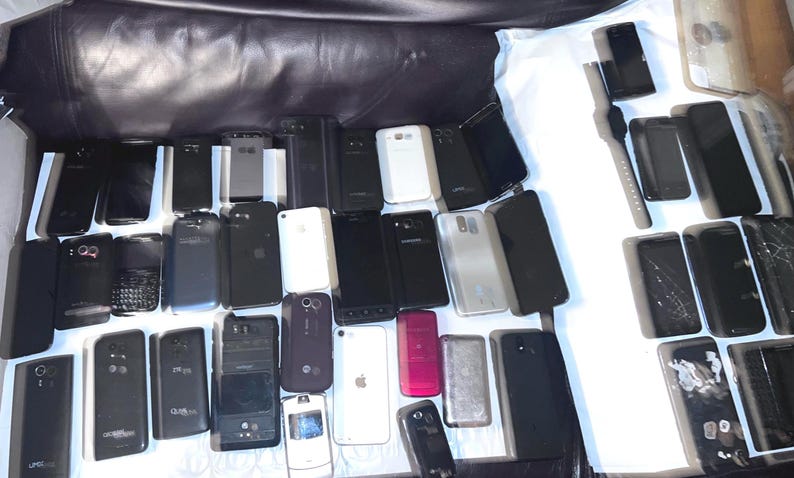 May include: A collection of various smartphones and a smartwatch, in different colors and designs. The phones are mostly black, with some white and other colored devices. Some phones have cracked screens.