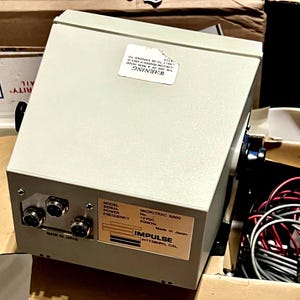 May include: A light gray electronic device labeled Impulse Microtrac 6800 with a 12VDC power rating and 200kHz frequency. The unit features three circular ports on the front panel, a warning sticker on the top, and a label indicating it is made in Japan.