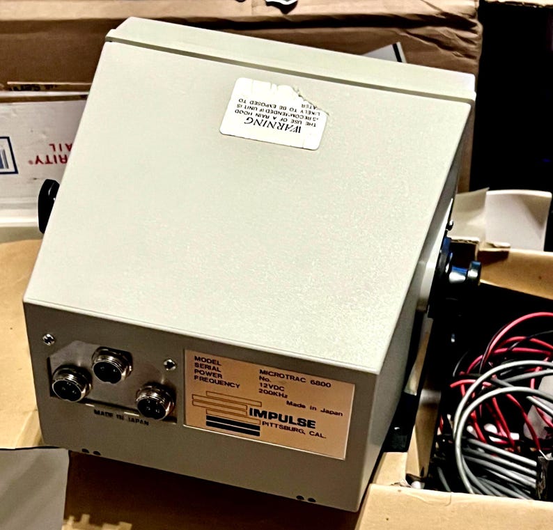 May include: A light gray electronic device labeled Impulse Microtrac 6800 with a 12VDC power rating and 200kHz frequency. The unit features three circular ports on the front panel, a warning sticker on the top, and a label indicating it is made in Japan.