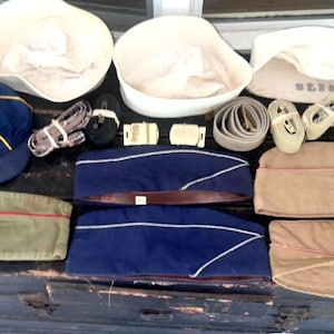 May include: A collection of vintage military hats, caps, belts, and accessories. Includes white sailor hats, blue and khaki caps, and a blue cap with a gold emblem. Also shown are belts and straps.