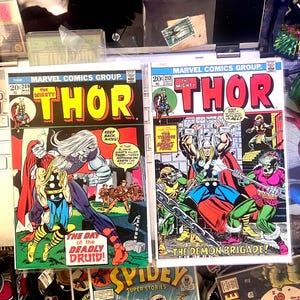 Puede incluir: Dos cómics vintage de Marvel Comics con Thor. Los cómics tienen portadas coloridas con escenas de acción y el título "Thor" en letras grandes y en negrita. Un cómic se titula "The Day of the Deadly Druid!" y el otro "The Demon Brigade!"