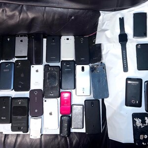 May include: A collection of various smartphones and a smartwatch, in different colors and designs, are arranged on a dark surface. The phones vary in size and shape, with some showing signs of wear. A few have visible logos.
