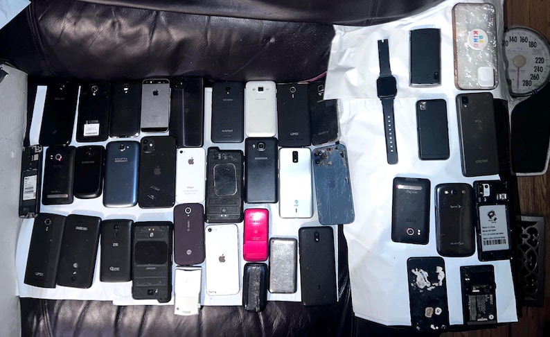 May include: A collection of various smartphones and a smartwatch, in different colors and designs, are arranged on a dark surface. The phones vary in size and shape, with some showing signs of wear. A few have visible logos.