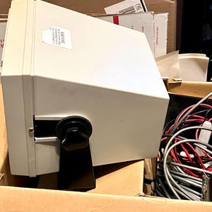 May include: A light gray electronic device with a trapezoidal housing, featuring a black adjustable mounting bracket secured by a circular knob on the side. A white rectangular warning label is affixed to the top surface of the unit.