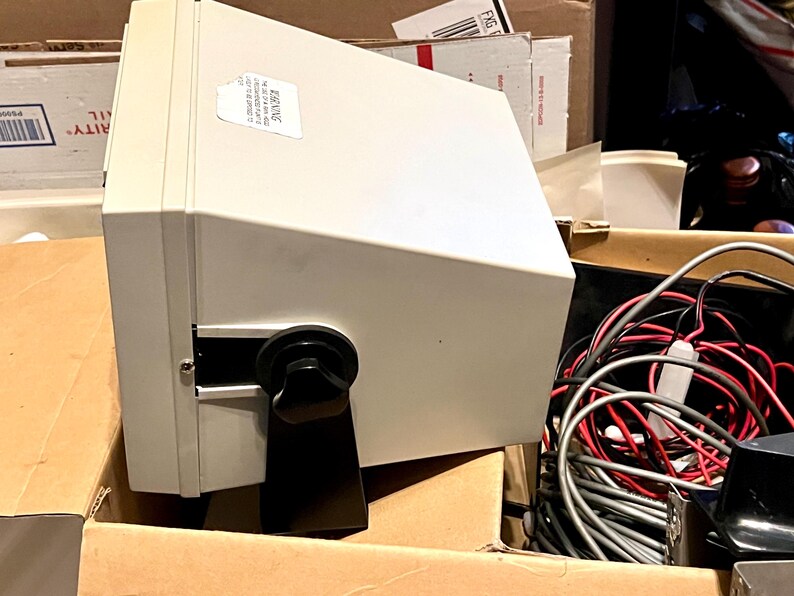 May include: A light gray electronic device with a trapezoidal housing, featuring a black adjustable mounting bracket secured by a circular knob on the side. A white rectangular warning label is affixed to the top surface of the unit.