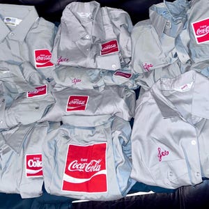 May include: A collection of light gray button-down shirts with red Coca-Cola logo patches. Some shirts have the name "Joe" embroidered in pink. The shirts are likely vintage or promotional items.