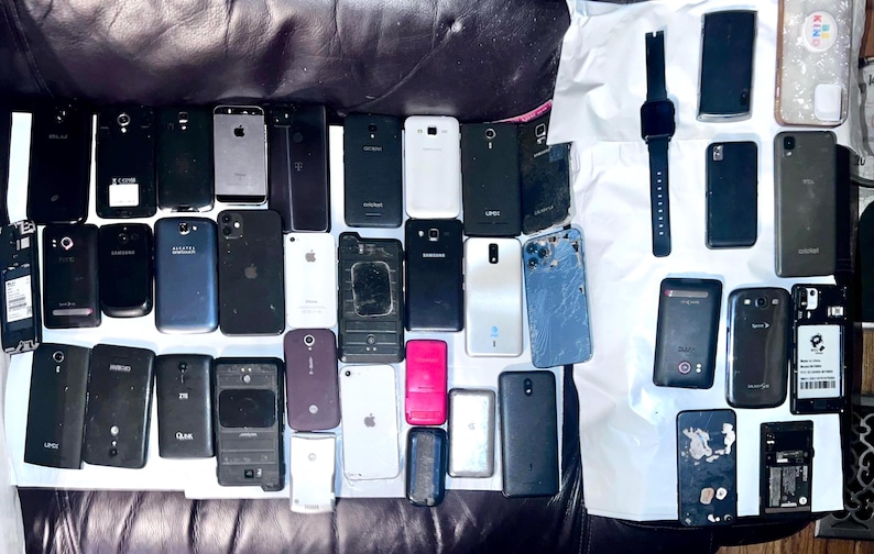 May include: A collection of smartphones and a smartwatch, in various colors and conditions, are arranged on a dark surface. The phones include models from different brands, with some showing signs of wear and tear. The smartwatch is black with a rectangular screen.