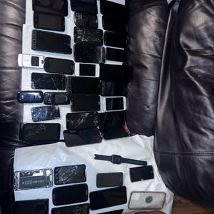 May include: A collection of various black smartphones, some with cracked screens, and a smartwatch are arranged on a white surface. The phones vary in size and design, suggesting a range of models. A scale is visible in the bottom right corner.