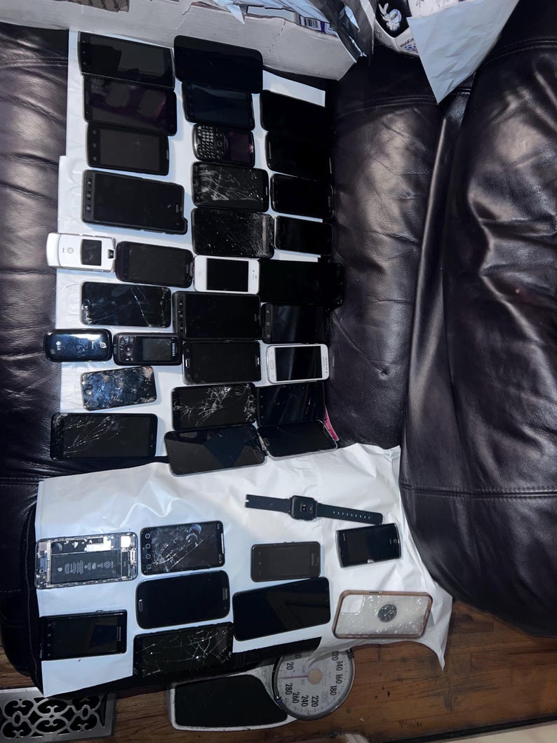 May include: A collection of various black smartphones, some with cracked screens, and a smartwatch are arranged on a white surface. The phones vary in size and design, suggesting a range of models. A scale is visible in the bottom right corner.