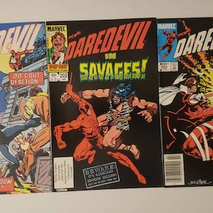 Puede incluir: Tres portadas de cómics con Daredevil, un superhéroe con un traje rojo y negro. Las portadas son de los números #201, #202 y #203. La primera portada muestra a Daredevil luchando contra una mujer con un traje negro. La segunda portada muestra a Daredevil luchando contra un hombre grande y salvaje. La tercera portada muestra a Daredevil atado por un hombre con un traje blanco.