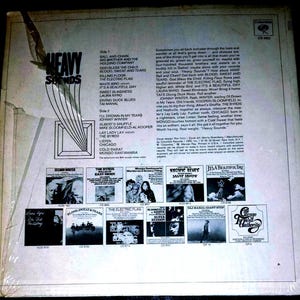 May include: The back cover of a Heavy Sounds vinyl record album featuring a tracklist, descriptive text, and small black and white thumbnail images of other Columbia record releases. The cover is white with black text and a torn plastic sleeve.