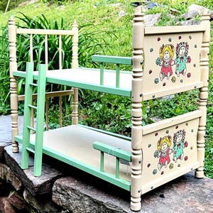 Op de afbeelding: Een poppenstapelbed in vintage-stijl met een crèmekleurig frame en mintgroene accenten. De hoofdborden tonen een kleurrijke illustratie van twee poppen. Het bed heeft een ladder en relingen voor het bovenste bed.