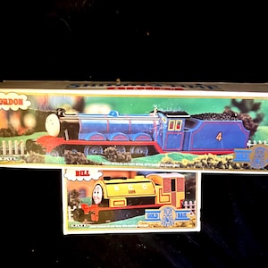 May include: Two Ertl die-cast metal toy train boxes. The top box features Gordon, a blue locomotive with the number 4. The bottom box features Bill, a yellow locomotive with the name Bill on the side. Both boxes display the Gold Rail logo.