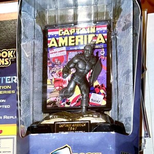 K&ouml;nnte beinhalten: Feine Zinnfigur von Captain America aus dem Goldenen Zeitalter, Teil der Comic Book Champions-Serie. Die Figur wird in einer transparenten Kunststoffbox mit einem Comic-Cover-Hintergrund pr&auml;sentiert. Die Verpackung enth&auml;lt das Marvel Comics-Logo.