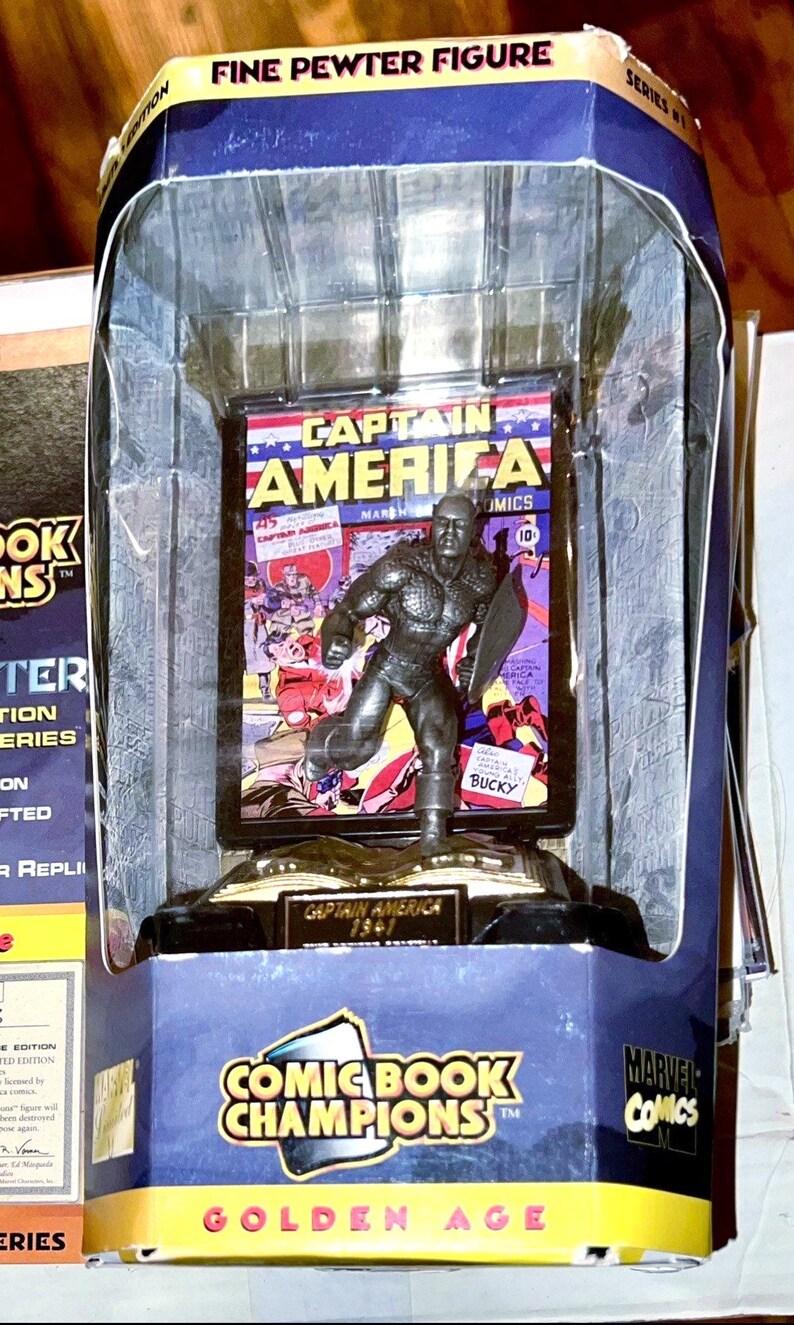 K&ouml;nnte beinhalten: Feine Zinnfigur von Captain America aus dem Goldenen Zeitalter, Teil der Comic Book Champions-Serie. Die Figur wird in einer transparenten Kunststoffbox mit einem Comic-Cover-Hintergrund pr&auml;sentiert. Die Verpackung enth&auml;lt das Marvel Comics-Logo.