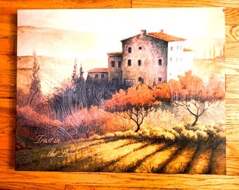 May include: A rectangular canvas print depicting a stone building nestled in a landscape with trees and fields. The artwork features warm orange and brown tones, with the text Trust in the Lord printed in the lower left corner of the scene.