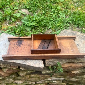 May include: A wooden tray with a rectangular box in the center. The tray and box are made of dark brown wood. The tray has a raised edge. The box has a slatted bottom. The tray is sitting on a stone wall.