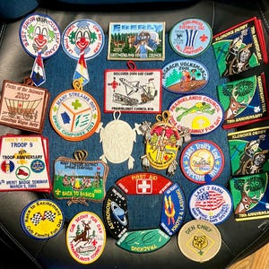 May include: A collection of various embroidered scouting patches featuring diverse shapes, colors, and designs, including text such as Best Show, Klondike Derby, and First Aid, arranged on a dark surface for display and historical documentation.