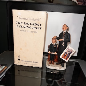 May include: A figurine of a man in a suit and a boy in a suit, holding a book that says "The Saturday Evening Post". The figurine is on a base with the words "Dave Grossman Designs" and "The Saturday Evening Post Co."