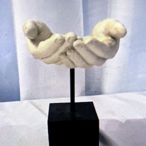 Puede incluir: Una escultura color crema de dos manos ahuecadas juntas, montada sobre un soporte de metal negro con una base cuadrada. Las manos parecen estar sosteniendo algo. La escultura está sobre un fondo blanco.