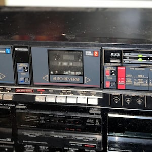 Puede incluir: Plataforma de casete estéreo Sharp negra, modelo RT-W600. Cuenta con dos reproductores de casete con función de inversión automática, un contador de cinta y varios botones de control. Debajo, una plataforma de casete doble estéreo Kenwood.