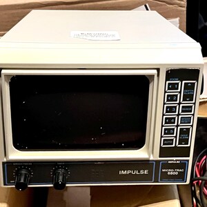 May include: An off-white Impulse Micro-Trac 6800 marine sonar display unit featuring a dark screen, a vertical control panel with numbered buttons, depth and range settings, and two black rotary knobs for brightness and gain control on the front.