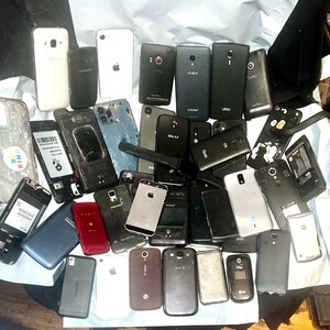 May include: A collection of various smartphones, some with cracked screens or missing components, in a variety of colors including black, white, and silver. The phones are scattered on a light-colored surface.