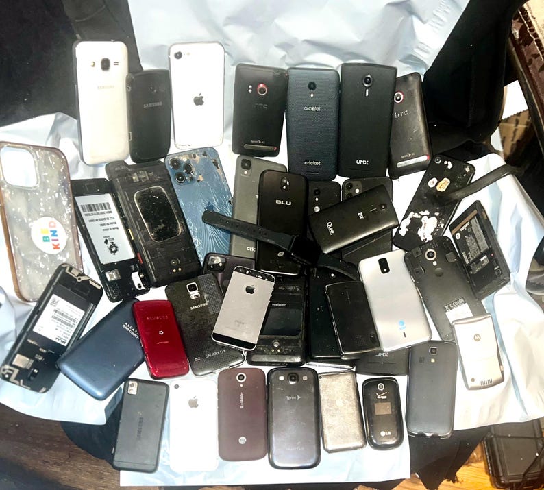 May include: A collection of various smartphones, some with cracked screens or missing components, in a variety of colors including black, white, and silver. The phones are scattered on a light-colored surface.