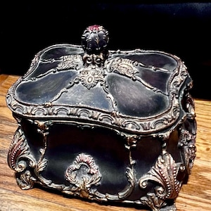 May include: A black and gold ornate jewelry box with a decorative lid and intricate details. The box has a raised design and a red jewel on the top.