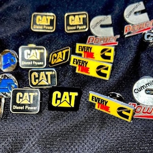 May include: A collection of metal pins and badges featuring various logos and text. The pins include the "CAT" logo in black and yellow, and "Cummins" logos in silver and red. Some pins have the text "Diesel Power" and "Every Time".