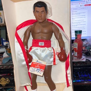 Puede incluir: Muñeca vintage de un boxeador en una caja de cartón. La muñeca lleva pantalones cortos blancos, un cinturón rojo con "EVERLAST", guantes rojos y botas blancas. Una capa roja y blanca está sobre los hombros de la muñeca. Una etiqueta dice "GREAT MOMENTS IN SPORTS."