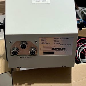 May include: A light gray electronic device with a metal casing, featuring three circular ports on the front panel. A label displays the text: MODEL MICROTRAC 6800, POWER 12VDC, FREQUENCY 200KHz, IMPULSE PITTSBURG, CAL., and MADE IN JAPAN.